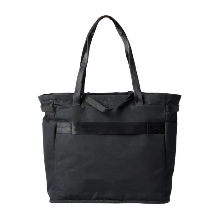 A black canvas tote bag with dual shoulder straps and a front utility pocket, hanging from a small gold wall hook against a white background.