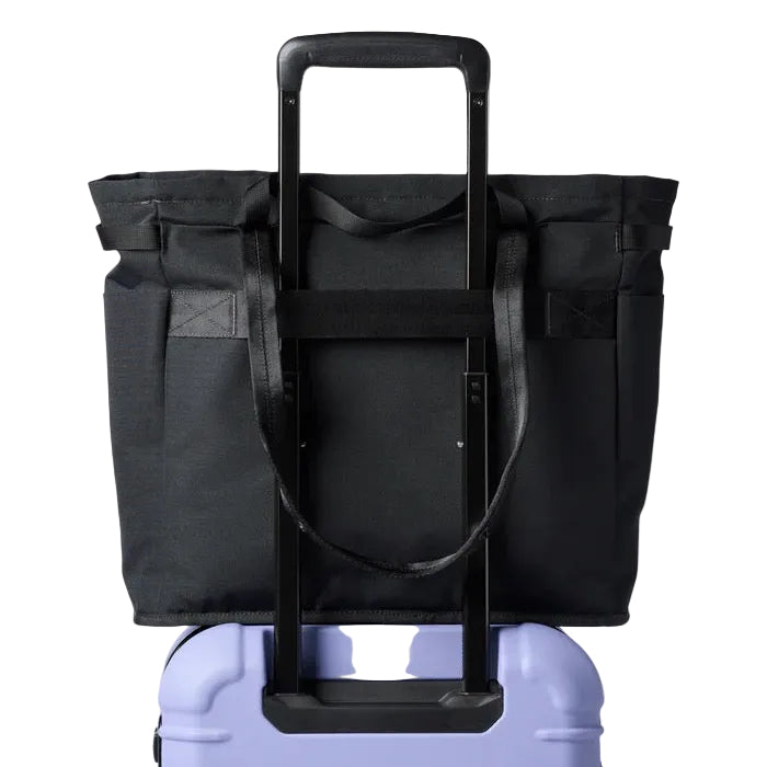 A black roll-top duffel bag with an extended black telescoping handle, shown resting on top of a lavender hard-shell suitcase against a plain white background.