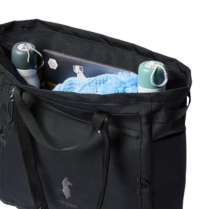 A black tote bag shown open, filled with a laptop, two water bottles, and a blue knit blanket, on a white background. The bag has a small deer logo and the brand name 'campmule' printed near the bottom front.